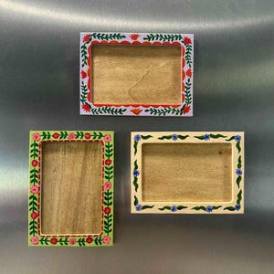 Puede incluir: Tres marcos rectangulares de madera con diseños florales. Uno tiene un fondo blanco con flores rojas y naranjas. Otro tiene un fondo verde con flores rosas. El tercero tiene un fondo crema con flores azules.