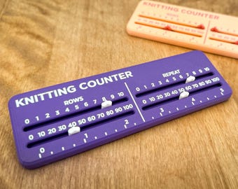 Può includere: Un contamaglie rettangolare viola e uno color pesca chiaro. Ogni dispositivo riporta la scritta KNITTING COUNTER e presenta due binari orizzontali per righe e ripetizioni, dotati di cursori bianchi scorrevoli per tenere il conteggio.