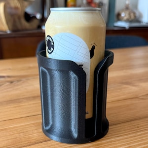 Pode incluir: Um porta-copos preto impresso em 3D com uma lata de cerveja dentro. A lata de cerveja é amarelo claro com um rótulo branco que diz "SPACE PUBLIC". O porta-copos está em uma mesa de madeira.