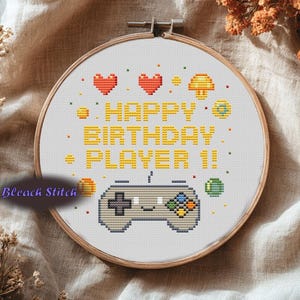 Op de afbeelding: Kruissteekborduurwerk in een houten hoepel met de tekst "HAPPY BIRTHDAY PLAYER 1!" en gepixelde harten, een paddenstoel en een gamecontroller. Het ontwerp is in geel, oranje, groen en grijs op een witte achtergrond.