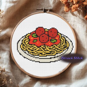 May include: Cross-stitch embroidery of spaghetti and meatballs on a white plate, set in a wooden embroidery hoop. The pasta is yellow, topped with red sauce and meatballs. The text "Bleach Stitch" is visible.