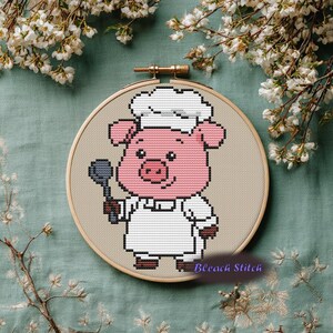 May include: Cross-stitch embroidery of a pink pig wearing a chef's hat and apron, holding a spoon. The pig is smiling. The embroidery is in a wooden hoop, with the text "Bleach Stitch" in the bottom right corner.