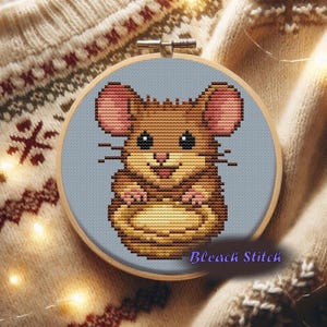 Może przedstawiać: Obręcz do haftu krzyżykowego z myszą z kreskówki z brązowym futrem, różowymi uszami i jasnoniebieskim tłem. Tekst "Bleach Stitch" jest napisany kursywą na dole. Obręcz jest osadzona w drewnianej ramie.