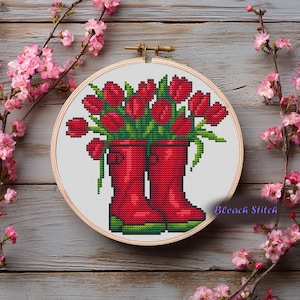 May include: Cross-stitch embroidery in a wooden hoop features red tulips in red rain boots. The embroidery is set against a white background and framed by a wooden hoop. The text "Bleach Stitch" is in the lower right corner.