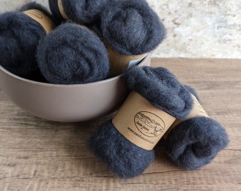 May include: A collection of dark gray wool roving, presented in a beige bowl and on a wooden surface. The wool is rolled into compact balls, some with brown paper labels. Ideal for fiber art and crafting projects.