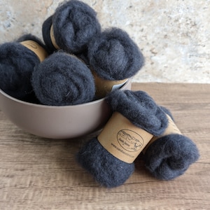 May include: A collection of dark gray wool roving, presented in a beige bowl and on a wooden surface. The wool is rolled into compact balls, some with brown paper labels. Ideal for fiber art and crafting projects.