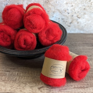 May include: A bowl filled with red wool roving, with two additional rolls of red wool roving on a wooden surface. The wool roving is packaged with a brown paper label that says "Santa Susana" and "www.santasusanaloft.com".