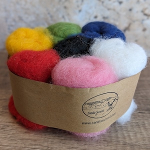 May include: A collection of colorful wool roving in a brown paper container. The wool is arranged in a circular pattern, with colors including yellow, red, green, black, pink, white, and blue. The container has a logo and website address printed on it.