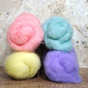 May include: Four colorful wool roving bundles in pastel shades. The colors include pink, yellow, teal, and purple. The wool is arranged in a stacked formation, with two bundles on top and two below. The background is a neutral, textured surface.