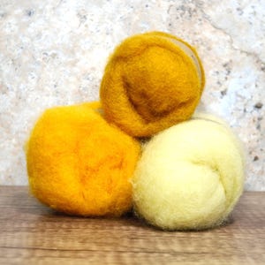 May include: Three balls of wool in shades of yellow and gold are stacked on a wooden surface. The wool balls have a soft, textured appearance, suggesting they are made for crafting or textile projects. The background is a mottled, neutral color.