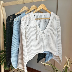 May include: Three crocheted sweaters hanging on wooden hangers. The sweaters are white, light blue, and black. The white sweater has a lace-up detail at the neckline.