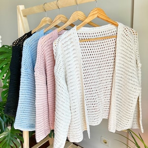 May include: Five crocheted cardigans hanging on wooden hangers. The cardigans are in black, light blue, pink, and two shades of white.