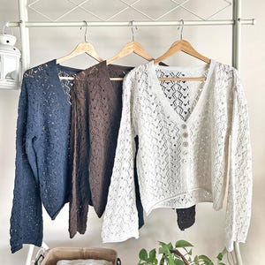 Peut inclure: Trois cardigans en maille ajourée, bleu marine, marron et blanc, sont suspendus à des cintres en bois. Le cardigan blanc est boutonné. Un portant en métal blanc soutient les cardigans, avec un panier de tissu plié et une plante en pot devant. Le panier porte l'inscription "sweet home".