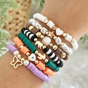 May include: A stack of colorful bracelets with various charms, including a butterfly, an evil eye, a star, a heart, and a bear. The bracelets are made of different materials, including beads, stones, and metal.