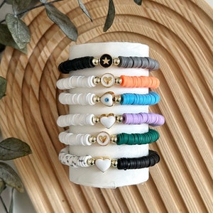 May include: A stack of colorful bracelets with white beads and gold charms. The charms include a star, an eye, and hearts. The bracelets are displayed on a white stand.