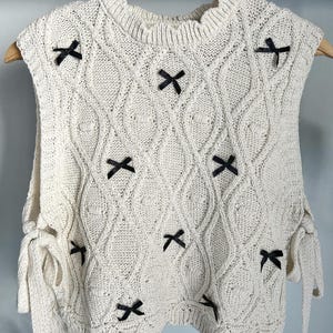 May include: Cream-colored knitted vest with a diamond pattern and black bow accents. The sleeveless top features tie details on the sides and is displayed on a wooden hanger. A stylish and unique addition to any wardrobe.