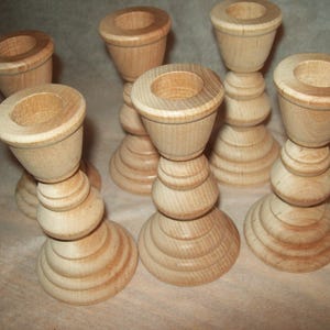 May include: A collection of seven unfinished wooden candle holders. Each holder features a round base, a series of stacked, rounded sections, and a top cup to hold a candle. The wood has a light, natural color.
