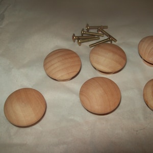 May include: Five round, unfinished light-colored wooden cabinet knobs with a smooth, domed surface and visible natural wood grain. A small cluster of five metallic, gold-toned screws is positioned near the top center of the arrangement.