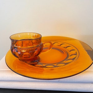 Può includere: Piatto da dessert in vetro ambrato con una tazza in vetro ambrato abbinata. Il piatto ha un bordo rialzato con un bordo smerlato e un motivo di piccoli cerchi. La tazza ha un bordo smerlato e un motivo di piccoli diamanti.