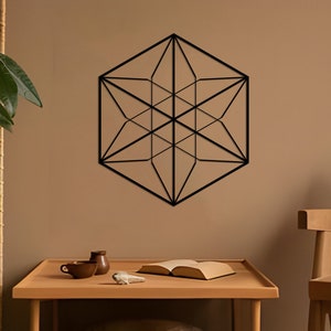 May include: A black geometric wall decor in the shape of a hexagon with a starburst design hangs on a light brown wall. A wooden table with a book and two ceramic cups sits in front of the decor. A wooden chair with a brown cushion is in the background.