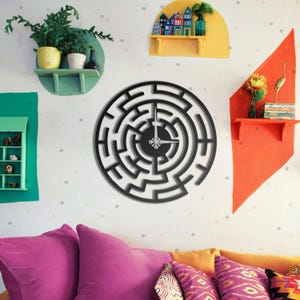 May include: A black, circular wall clock with a maze-like design. The clock hands are silver. The clock is mounted on a white wall with colorful shelves and decorative items. The room has a modern, artistic aesthetic.