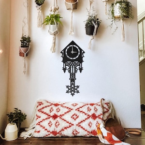 May include: A black metal cuckoo clock with a decorative design is mounted on a white wall. The clock is surrounded by hanging planters with greenery. A patterned cushion and other decor are below the clock.