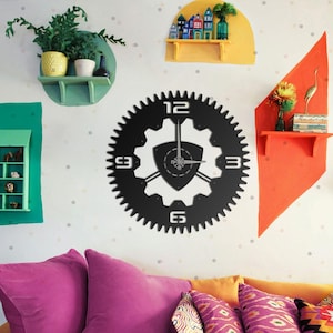 May include: A black metal wall clock with a gear design. The clock features large white numbers at the 12, 3, 6, and 9 o'clock positions. The clock is mounted on a white wall with colorful decorative shelves and pillows.