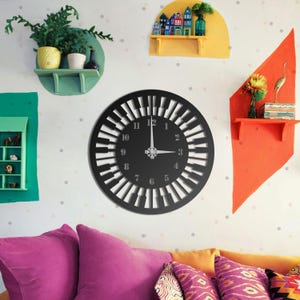May include: A black wall clock with a piano key design. The clock has silver hands and numbers. The clock is mounted on a white wall with colorful shelves and decorative items.