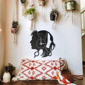 Puede incluir: Reloj de pared de metal negro con las siluetas de un hombre y una mujer, con una esfera de reloj en el centro. El diseño incorpora tijeras, lo que sugiere un tema de salón de belleza o peluquería. El reloj está montado en una pared blanca.