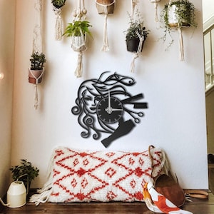 Puede incluir: Reloj de pared de metal negro con la cara de una mujer con cabello suelto, una esfera de reloj, tijeras y un peine. El reloj está montado en una pared blanca, con plantas colgantes y un cojín decorativo debajo. Un artículo de decoración único para salón o spa.