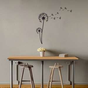 Puede incluir: Una pared gris claro con una calcomanía de pared de diente de león negro. La calcomanía presenta un diente de león grande con semillas que se van volando con el viento. Hay una mesa de madera con dos taburetes de madera frente a la pared.
