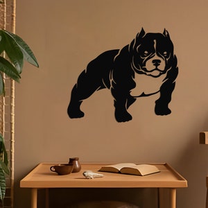 May include: Black silhouette of a muscular American Bully dog breed standing on a light brown wall. The dog is facing forward with its head slightly tilted. The background includes a wooden chair with a brown cushion and a small table with two mugs and a book.