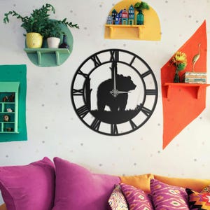 Puede incluir: Reloj de pared de metal negro con una silueta de oso dentro de un marco circular con números romanos. Las manecillas del reloj son visibles y el oso está sobre hierba. El reloj está montado en una pared blanca con decoración colorida.