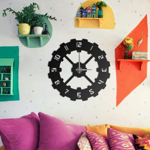 Peut inclure: Une horloge murale noire en forme d'engrenage avec des chiffres et des aiguilles blancs. L'horloge est fixée sur un mur blanc avec des étagères colorées et des objets décoratifs. Le design de l'horloge ajoute une touche industrielle moderne à la pièce.
