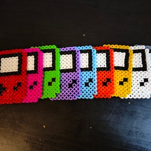 May include: A row of eight colourful pixel art Game Boy handhelds made with perler beads. The Game Boys are in red, pink, green, purple, blue, orange, yellow, and grey.
