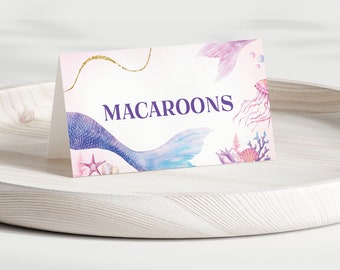 May include: A folded card with the word "MACAROONS" in purple text. The card features a mermaid tail, jellyfish, and seashell illustrations in shades of blue, pink, and purple. A gold glitter accent is also present.