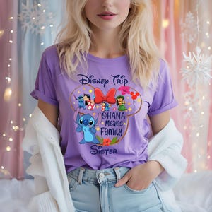 May include: A lilac-colored t-shirt featuring a Disney-themed graphic. The design includes the text "Disney Trip" and "Ohana Means Family Sister" along with cartoon characters and a red bow. The shirt has rolled-up sleeves.