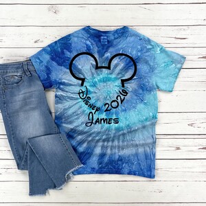 Pu&ograve; includere: Una maglietta blu tie-dye con un design a testa di Topolino nero e la scritta "Disney 2026 James". La maglietta &egrave; abbinata a un paio di jeans blu.