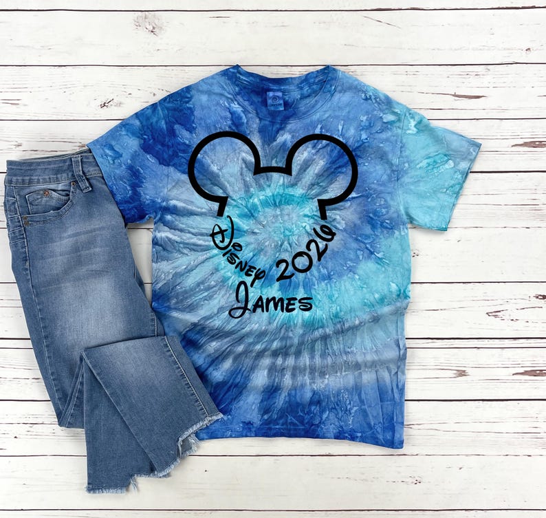 Pu&ograve; includere: Una maglietta blu tie-dye con un design a testa di Topolino nero e la scritta "Disney 2026 James". La maglietta &egrave; abbinata a un paio di jeans blu.