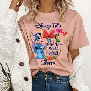 May include: A light pink t-shirt featuring a Disney-themed graphic. The design includes the text "Disney Trip" and "Ohana Means Family" with images of Stitch and other characters. A red bow and floral accents complete the design.