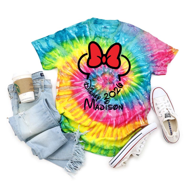 Pu&ograve; includere: Una maglietta tie-dye con un design di Minnie Mouse, jeans, una tazza di caff&egrave; e scarpe da ginnastica bianche. La maglietta ha un fiocco rosso e il testo "Disney 2020 Madison". La maglietta &egrave; un vortice di colori tra cui blu, giallo e rosa.