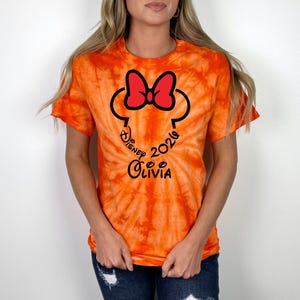 Pu&ograve; includere: T-shirt arancione tie-dye con un design di Minnie Mouse nero e rosso. Il design include il testo "Disney 2024 Olivia" in un carattere corsivo. La maglietta ha maniche corte e girocollo.