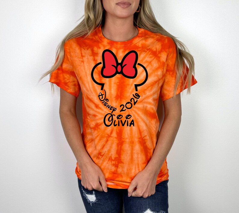 Pu&ograve; includere: T-shirt arancione tie-dye con un design di Minnie Mouse nero e rosso. Il design include il testo "Disney 2024 Olivia" in un carattere corsivo. La maglietta ha maniche corte e girocollo.