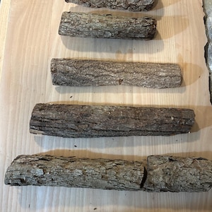 May include: Five cylindrical pieces of natural wood with rough, textured brown bark arranged in a horizontal stack. The logs vary in length and thickness, displaying organic patterns, knots, and fissures across their surfaces.