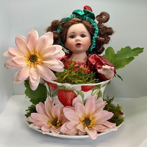 May include: A porcelain doll with curly brown hair and a green headband sits inside a white teacup decorated with red strawberries. The teacup is filled with green moss and surrounded by large, pale pink flowers on a matching saucer.