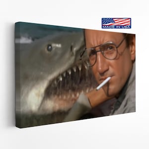 May include: A canvas print featuring a scene from the movie "Jaws." The image shows a close-up of a shark's open mouth with teeth, and a man wearing glasses and holding a cigarette. The print has a "Made in USA" label.