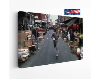 May include: A rectangular canvas print featuring a scene of a person running down a city street lined with market stalls and cardboard boxes. The runner wears a grey long-sleeved shirt, grey trousers, and a red headband. A small MADE IN USA logo is visible.