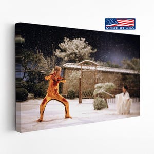 May include: A canvas print depicting a scene from a movie, featuring a person in a yellow jumpsuit wielding a sword in a snowy outdoor setting. The image includes a small "Made in USA" flag.
