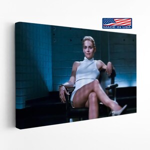 May include: A rectangular canvas wall art print featuring a scene from the film Basic Instinct. The image shows a person with blonde hair wearing a white sleeveless mock neck dress, sitting in a black chair with legs crossed in a blue-toned room.