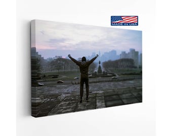 May include: A rectangular canvas print featuring a person standing with arms raised in victory on stone steps overlooking a city park at dawn. The scene includes a distant skyline, trees, and a road. A small logo in the corner reads MADE IN USA.